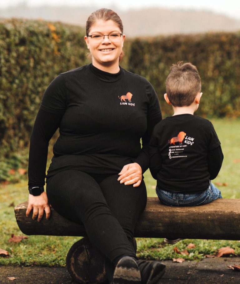 Helen Häge, Resilienztrainerin von Lion Kids, sitzt lächelnd auf einer Bank. Ein kleiner Junge mit dem Rücken zur Kamera sitzt neben ihr.