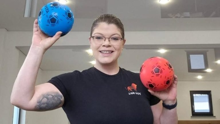 Helen Häge hält lächelnd einen blauen und einen roten Ball in einem Fitnessraum.