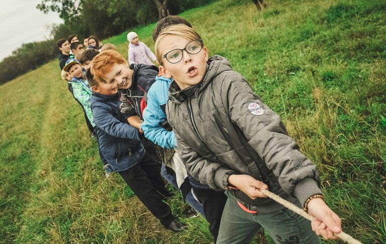 Kinder ziehen gemeinsam an einem Seil in einer Wiese, umgeben von Bäumen.
