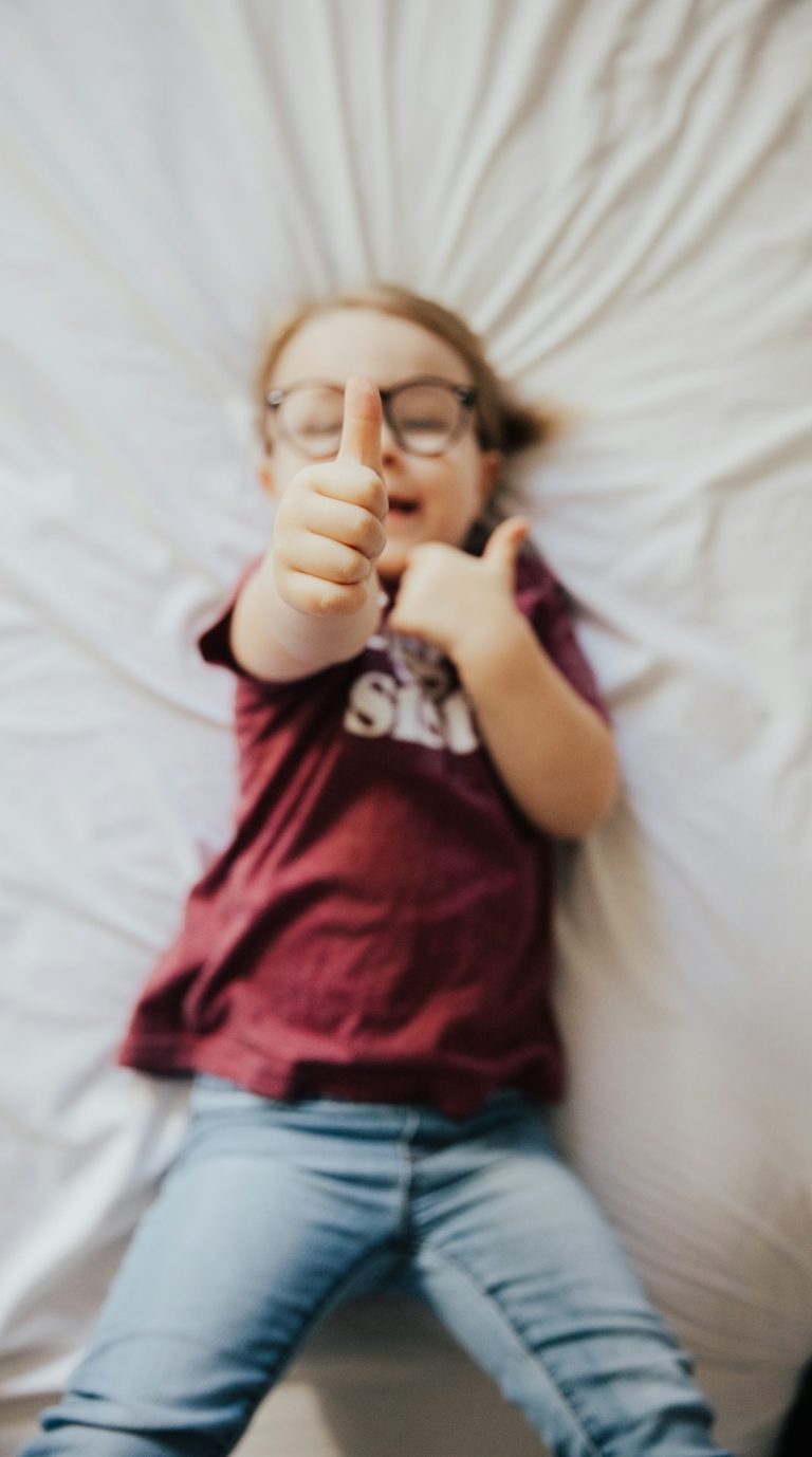 Ein Kind liegt auf einem Bett und zeigt mit beiden Händen Daumen hoch.