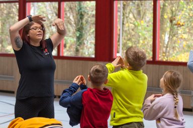 Die Resilienztrainerin Helen Häge zeigt einer Gruppe von Kindern Bewegungen in einem Raum mit großen Fenstern.