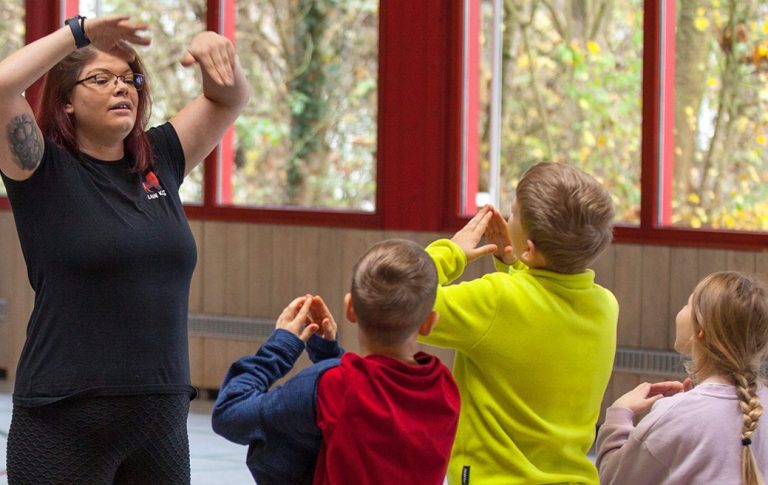 Die Resilienztrainerin Helen Häge zeigt einer Gruppe von Kindern Bewegungen in einem Raum mit großen Fenstern.